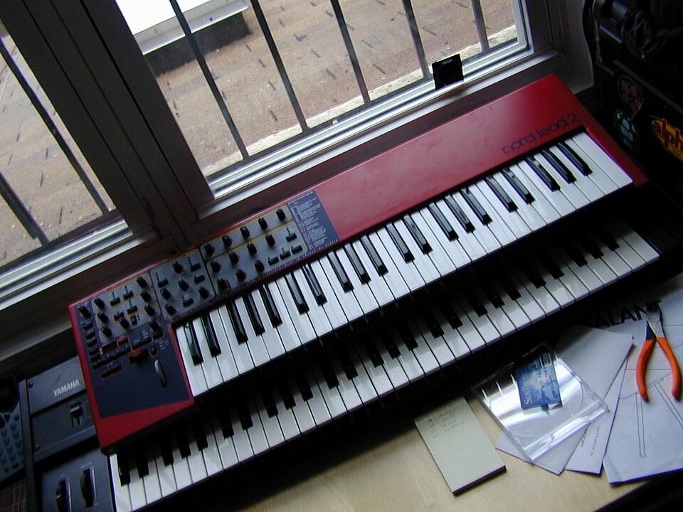 A red Clavia Nord Lead 2 keyboard on top of a Yamaha DX7 by a window.