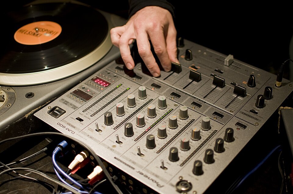 A silver Pioneer DJM-600 DJ mixer next to a turntable, with a hand on a fader.