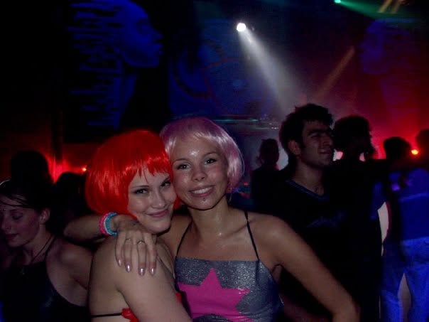 Two women in red and pink wigs in the crowd, painted faces on the backdrops behind them.