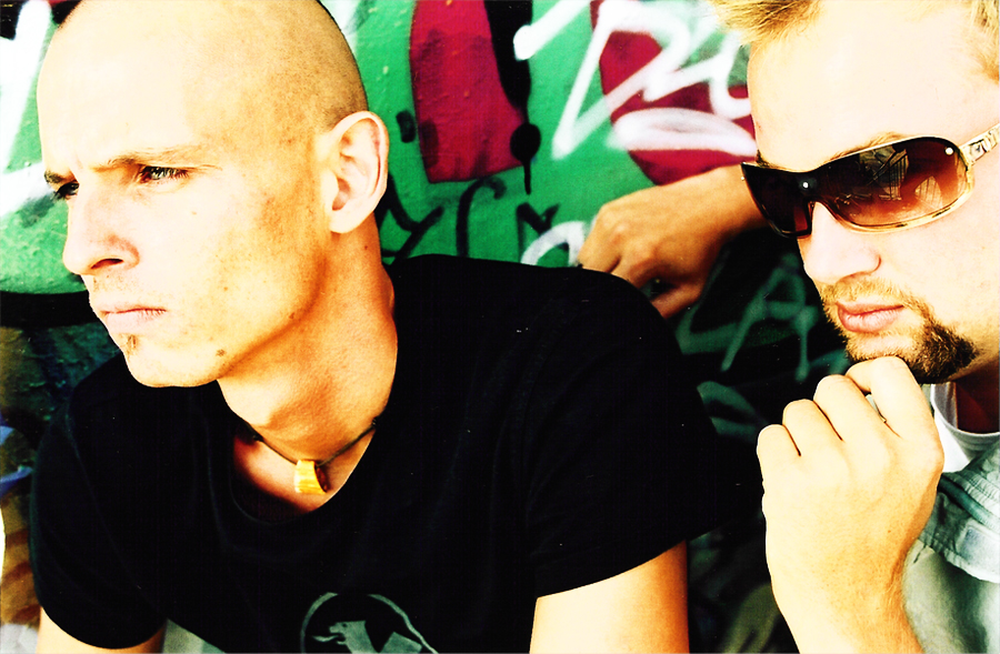 The two members close up in front of a graffiti wall: one with a shaved head and a black T-shirt looking to the side, the other in sunglasses with his chin on his hand.