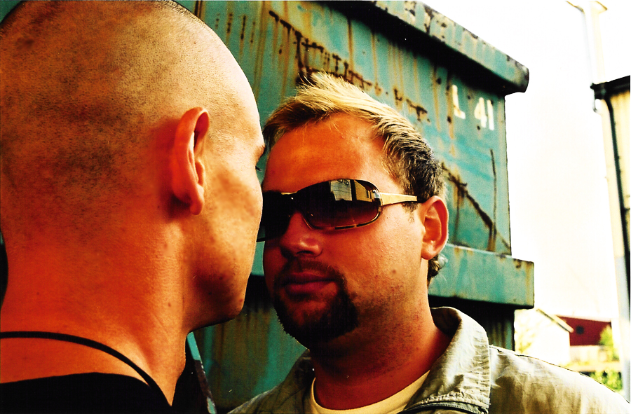 The two face to face by a green container; the one in sunglasses looks into the camera.