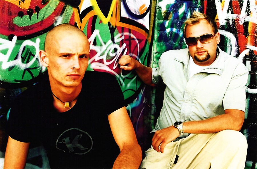 Both sitting against a graffiti wall, one in a black T-shirt, the other in a light shirt and sunglasses.