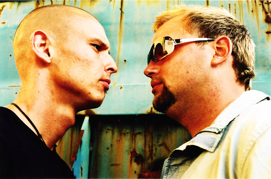 The two in profile, face to face in front of a rusty container.