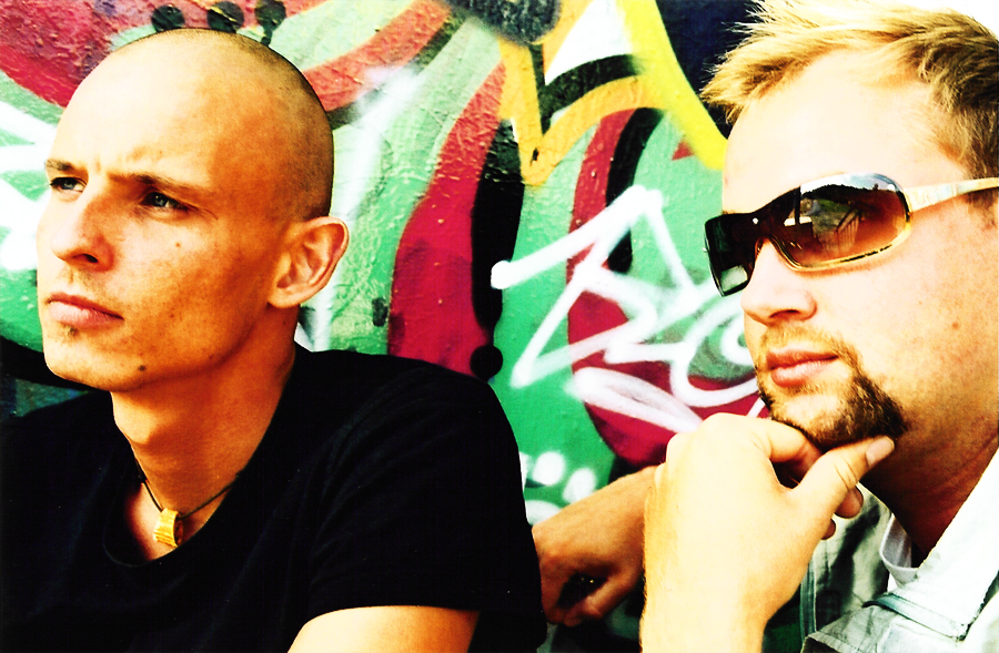 Close-up of both in front of the graffiti wall, looking to the left.