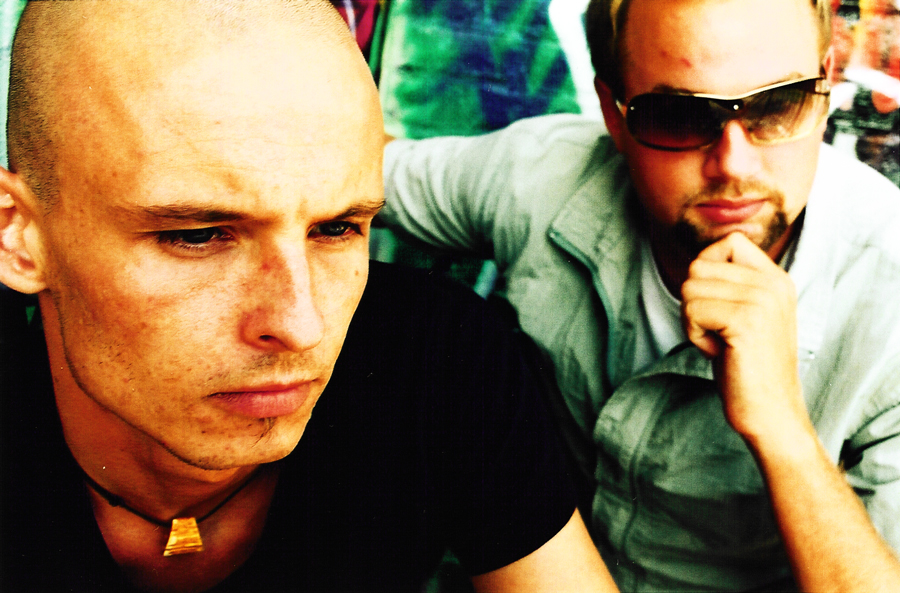 One member looking down in the foreground, the other behind him in sunglasses, chin on his hand.