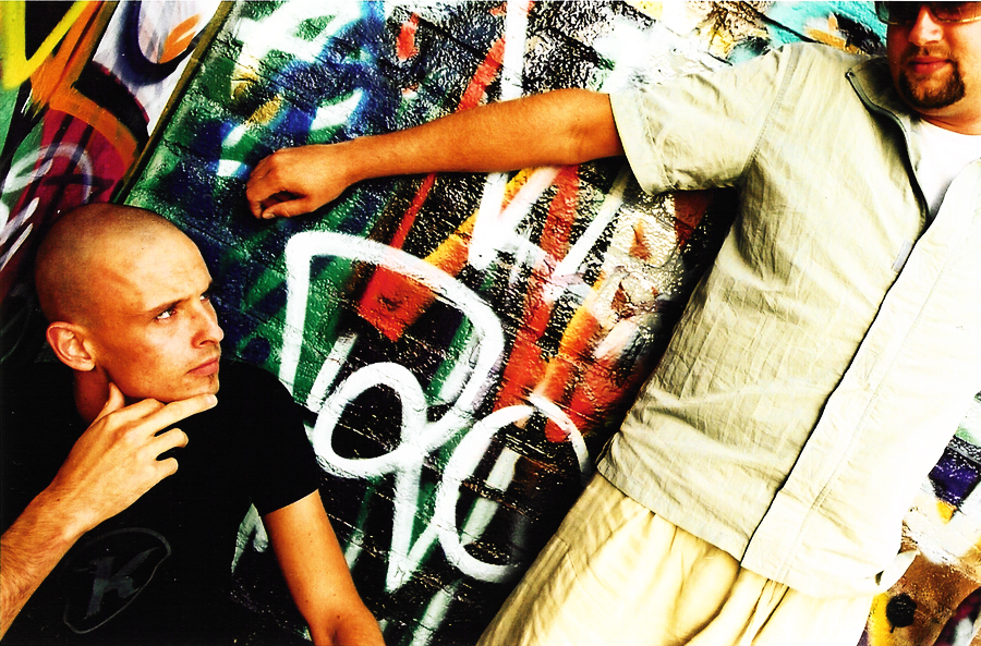One member crouching with his chin on his hand, the other leaning on the graffiti wall above him.