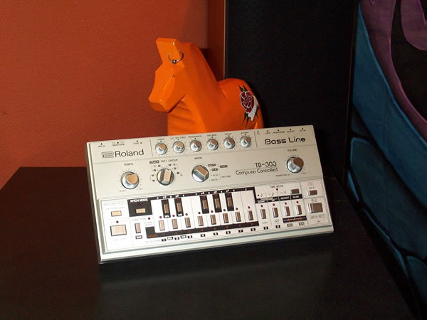 A Roland TB-303 Bass Line on a black surface, an orange Dala horse behind it.