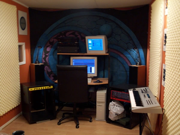 A small studio room: grey egg-crate foam on the walls, a painted blue and purple circular backdrop behind a desk with two computer monitors, a black office chair, a rack with a red synthesizer module and a white keyboard on a stand.