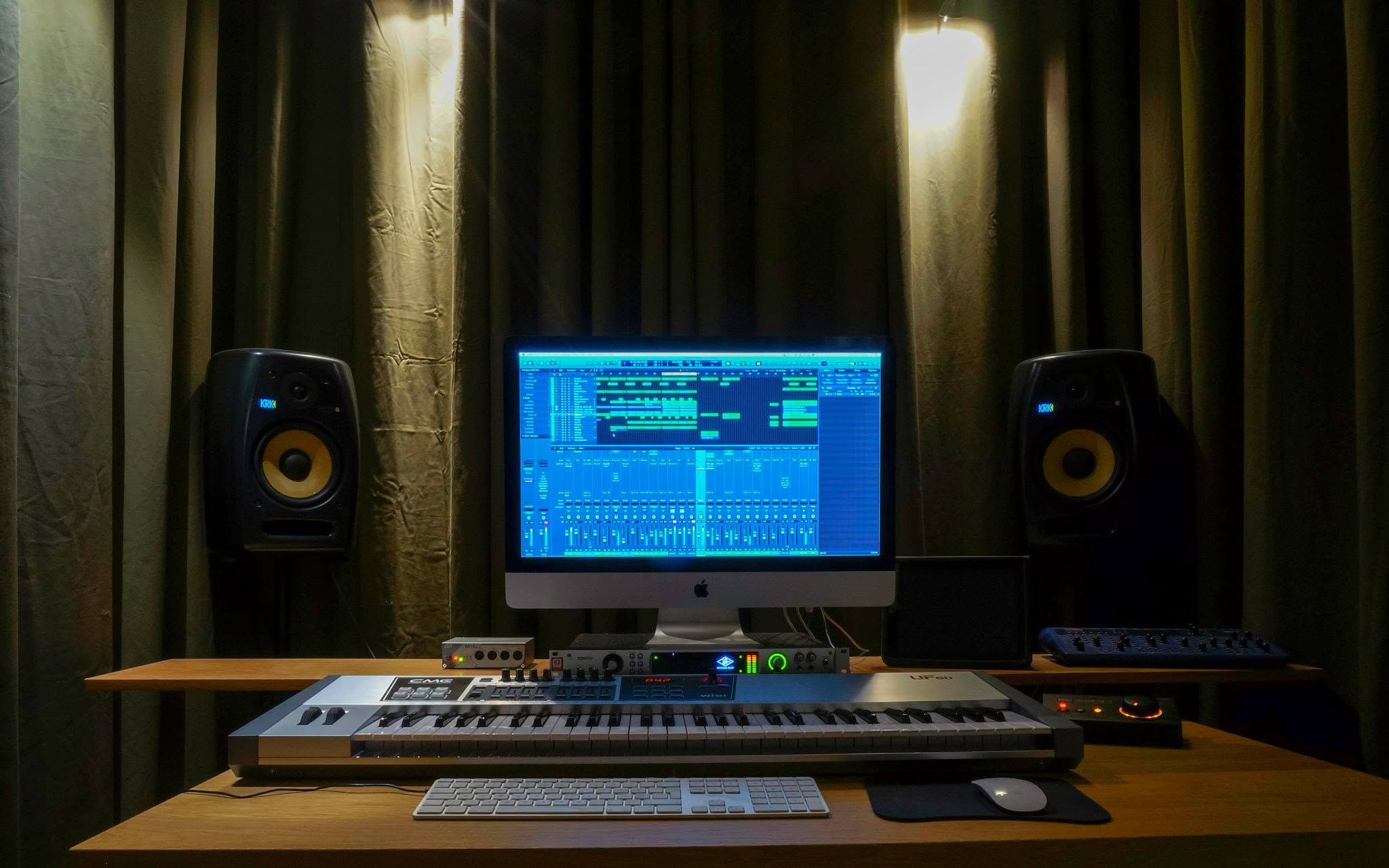 The studio desk between green curtains: two KRK monitors, an iMac with an arrangement on screen, a CME UF60 keyboard, a Universal Audio Apollo interface and the blue Novation Nova on the shelf.