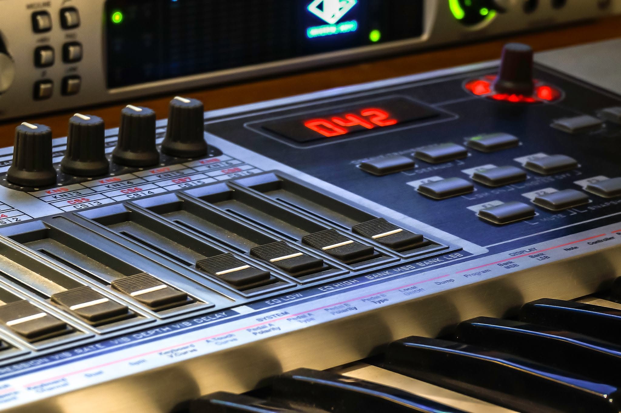 Close-up of the keyboard's faders, buttons and red display, with rack equipment behind it.