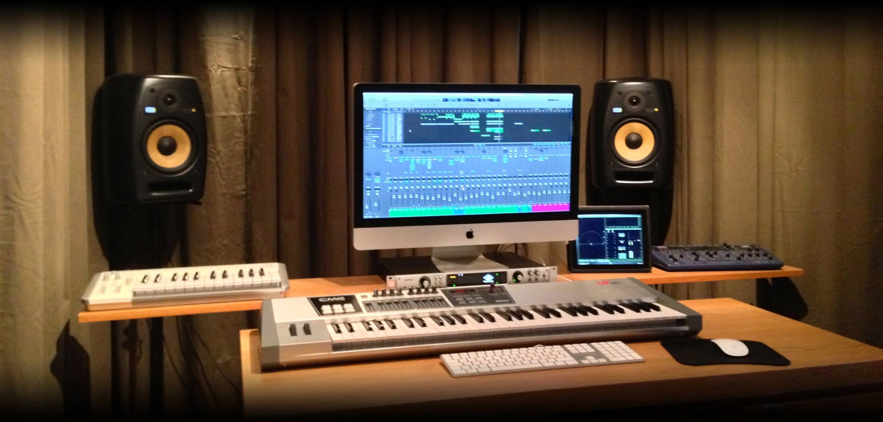 The studio desk lit by two spotlights between curtains: KRK monitors either side of an iMac, with a keyboard and a synthesizer on the desk.