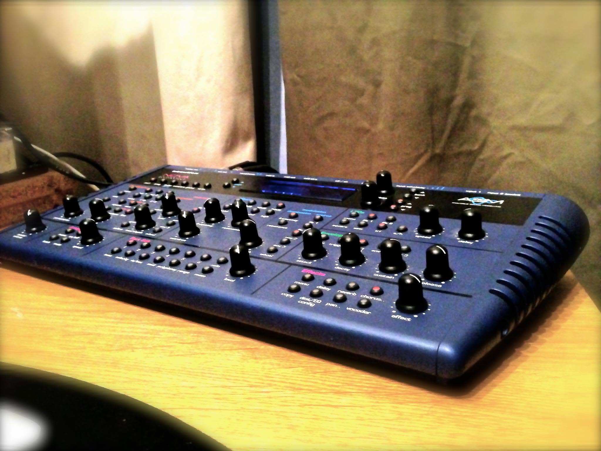 The blue Novation Nova synthesizer with its many knobs on a wooden desk in warm light.